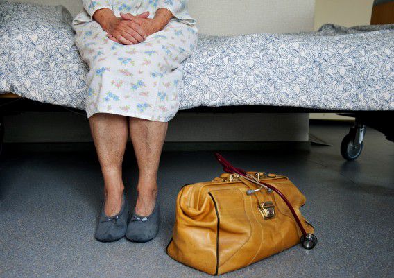 Een bedlegerige oudere dame in nachtpon zit op bed tijdens doktersbezoek in een aanleunwoning.