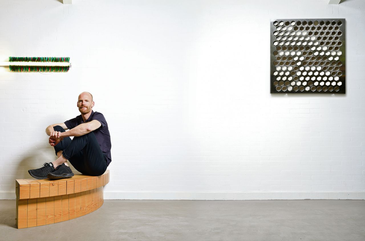 Chris Kabel zit op een deel van zijn Wood Ring (2010), een cirkelvormige bank gemaakt uit een balk hout. Boven hem hangt Office Party (2014): door aan een touwtje te trekken verandert een kille tl-lamp in feestverlichting.