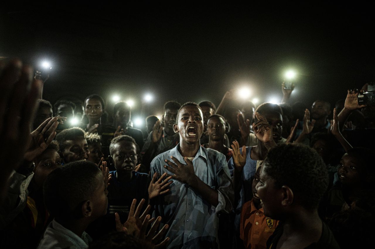 Genomineerd voor World Press Foto van het Jaar en Algemeen Nieuws - Enkel. Dictator Bashir Soedan werd afgezet, maar de demonstraties voor een democratischer systeem duurden daarna voort. Op deze foto draagt een jongen een gedicht voor te midden van een groep jonge demonstranten. De elektriciteit is afgesloten, maar hun gezichten worden verlicht door het schijnsel van hun mobiele telefoons.