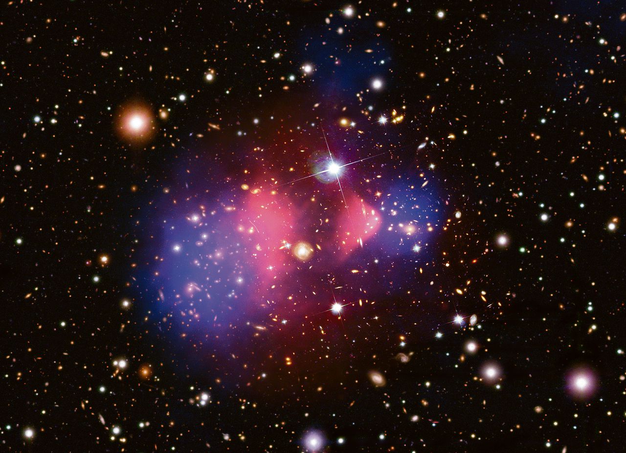 het Bullet-Cluster, waarin twee groepen van sterrenstelsels op elkaar botsen, 3,4 miljard lichtjaar van de aarde. De paarse wolk (in röntgenlicht gefotografeerd) is super heet gas. Uit de relatie tussen sterrenstelsels en het gas kan het bestaan van ‘donkere materie’ worden afgeleid.