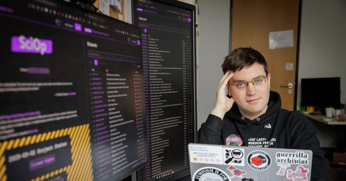 Henrik Schönemann sitting in front of a desk with multiple computer monitors and a laptop. The laptop is adorned with various stickers, including one that reads 'guerrilla archivist', one with 'Protect Trans Lives', another that says 'Feminism is for everyone.' He is resting his head on their hand be in contemplative pose. The monitors display a dark-themed interface with purple accents, showing a web application with the title 'SciOp.' The left monitor shows a section titled 'Details,' while the right monitor displays a list of datasets.