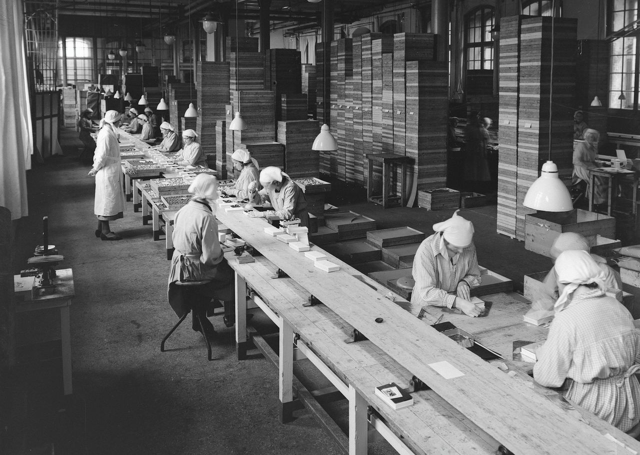 Fabriekshal waar vrouwen deels via een lopende band dozen vullen met bonbons die in grote houten dozen zitten. Weesp, Noord-Holland, 1910.