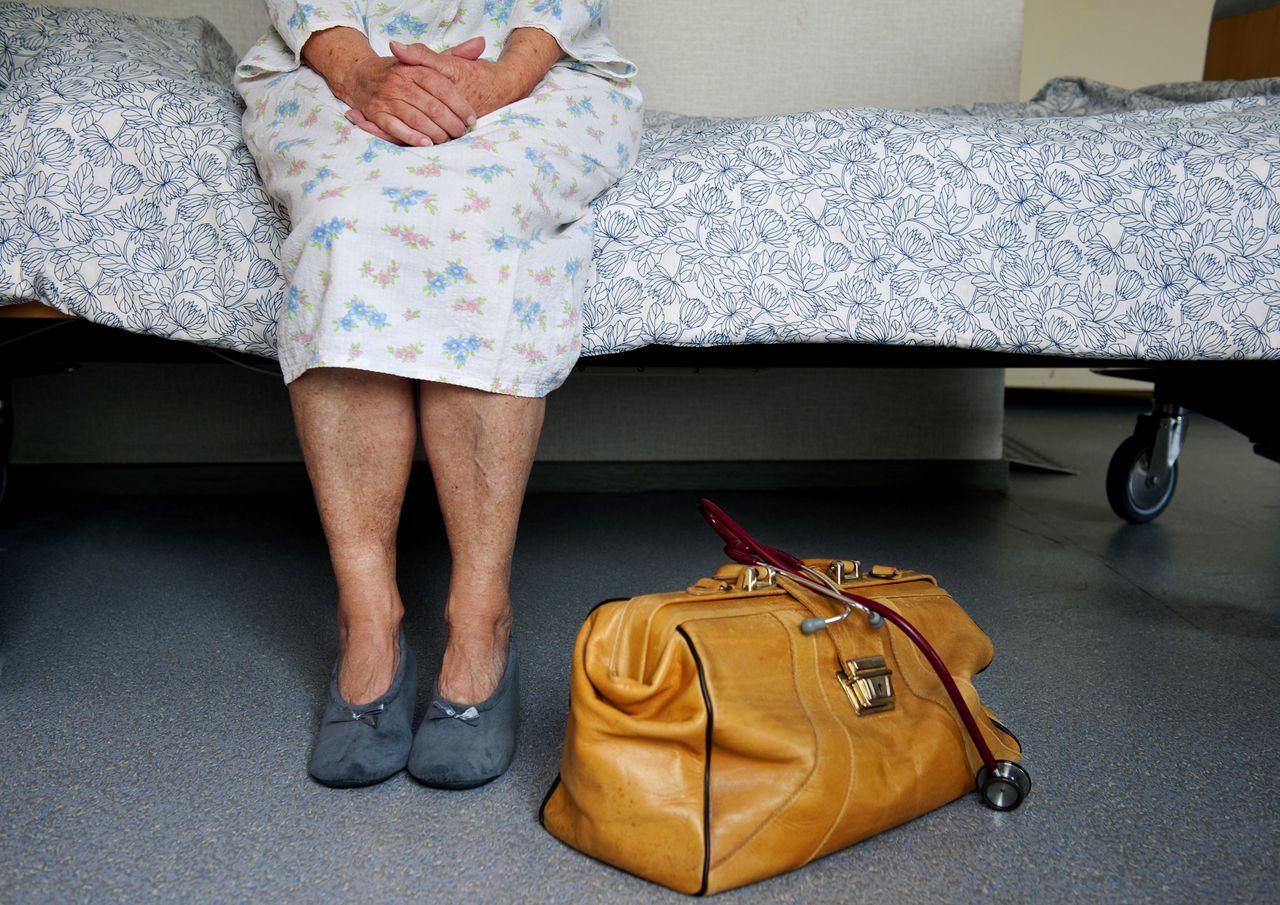 Een oudere dame in nachtpon zit op bed tijdens doktersbezoek.