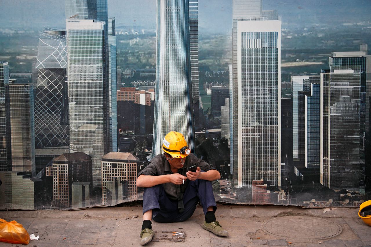 Een bouwvakker pauzeert op een bouwplaats. Op de achtergrond een poster met de wolkenkrabbers van het zakelijke centrum van Beijing.
