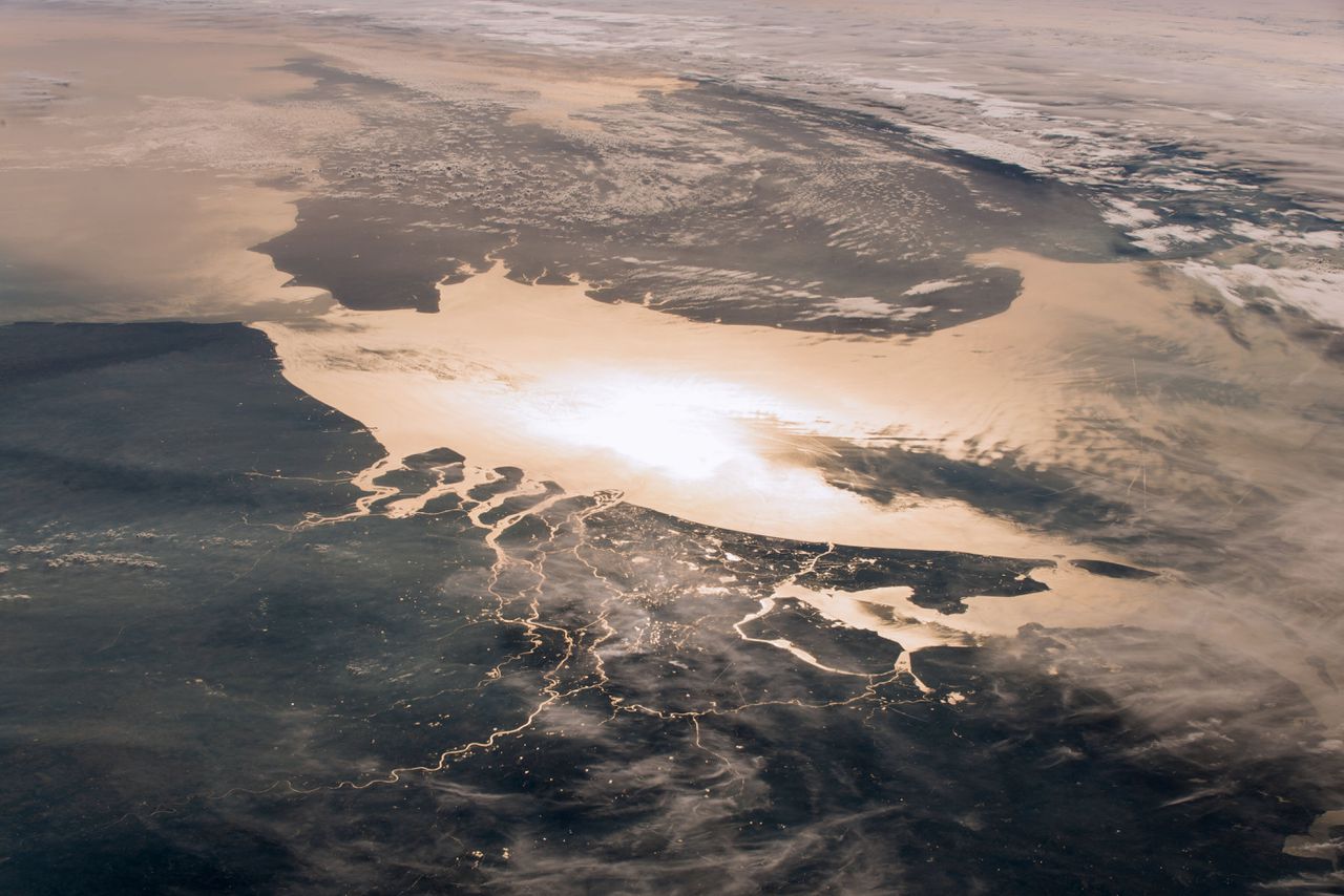 Nederland gezien vanuit het ruimtestation ISS. In de reflectie van het zonlicht tekent de rivierendelta zich scherp af tegen het donkere land.