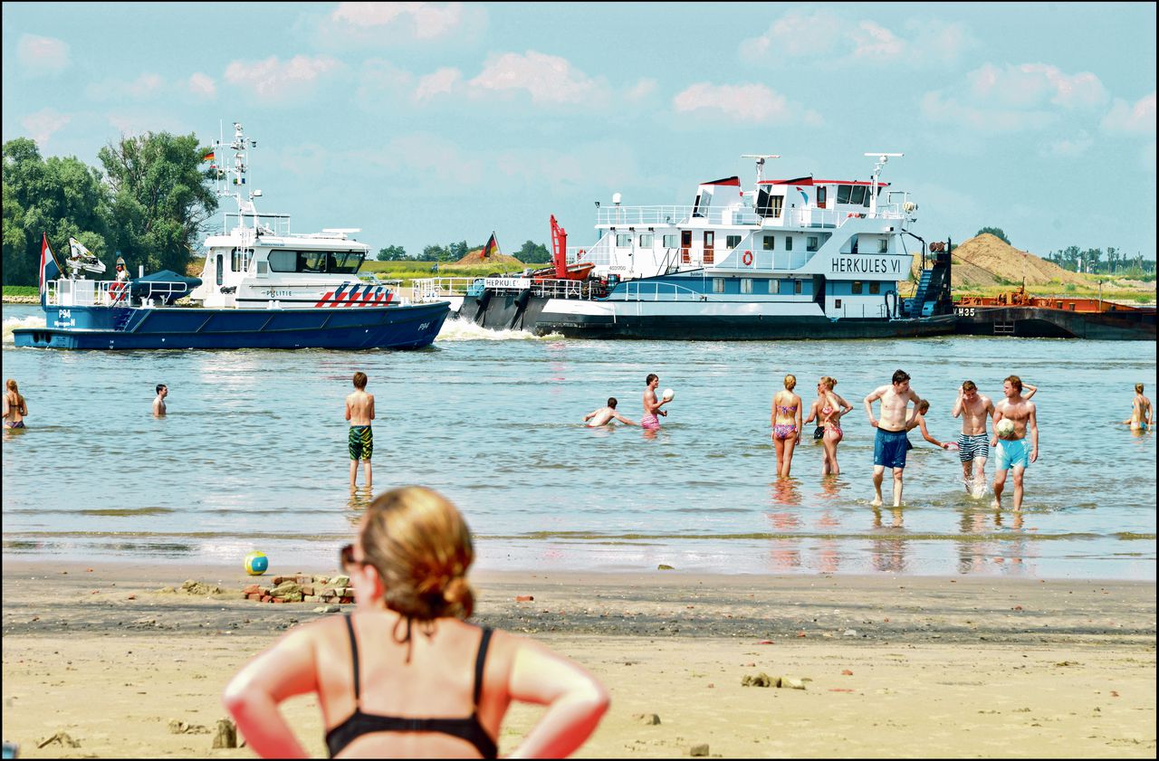 Een zomerse dag op het Waalstrand van Nijmegen in de Ooijpolder. Het zwemmen in de rivier is gevaarlijk vanwege de verraderlijke stroming en de drukke scheepvaart. Als een grote zesbakscombinatie langs Nijmegen vaart, begeleidt de politieboot deze langs de strandjes om te zorgen dat er geen zwemmers te dicht bij het schroefwater komen. Foto Flip Franssen