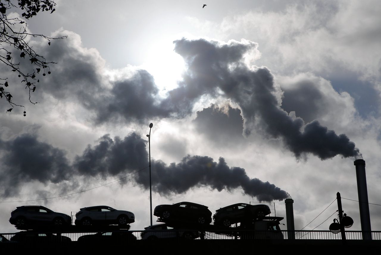 Een truck met auto's rijdt voor een fabriek die rookpluimen uitstoot.